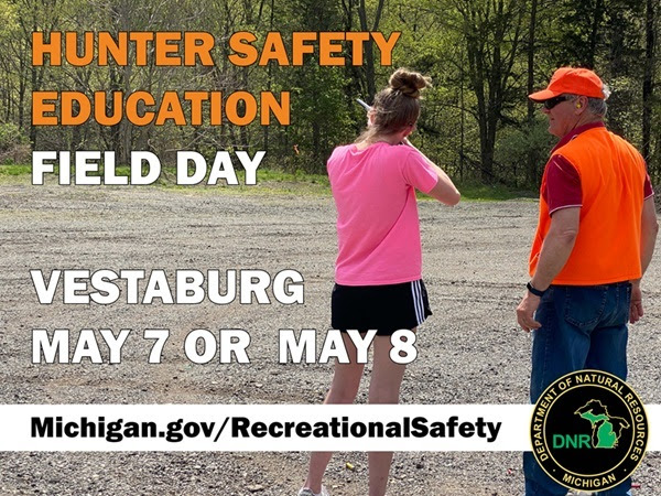 woman in pink T-shirt and black shorts aims a rifle toward a forested area as a man in hunter orange clothing looks on