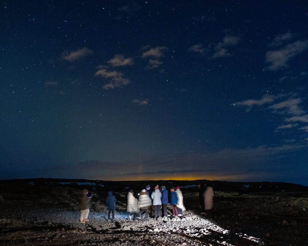 A group of tourists stand in the middle of an empty field. They are illuminated by headlights. The sky above them is blue and vast, with a shimmer of orange at the horizon.