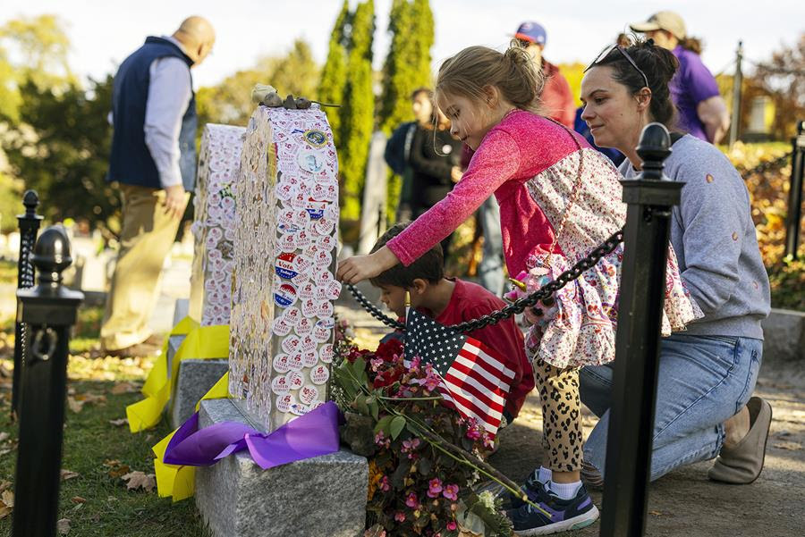 A little girl with fair skin and dirty blond hair puts an 'I Voted' sticker on a gravestone covered in other 'I Voted' stickers. There is a little boy crouched beside her looking at the gravestone beside it. There is an older white woman with dark hair standing behind the two children.