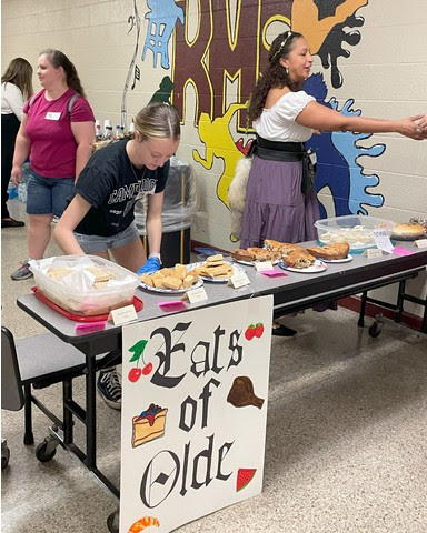 Students work at a food table, a banner reads Eats of Olde