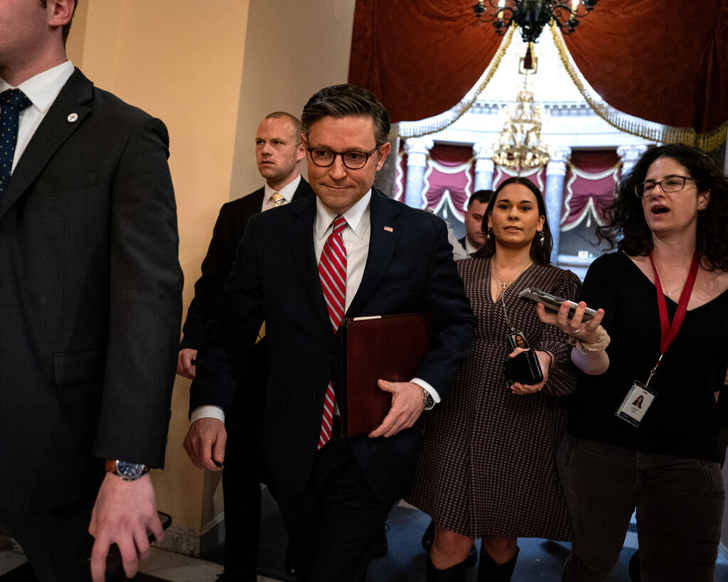 House Speaker Mike Johnson, wearing a dark suit and holding a portfolio under his left arm, looks down as he walk through the Capitol as other people follow him.