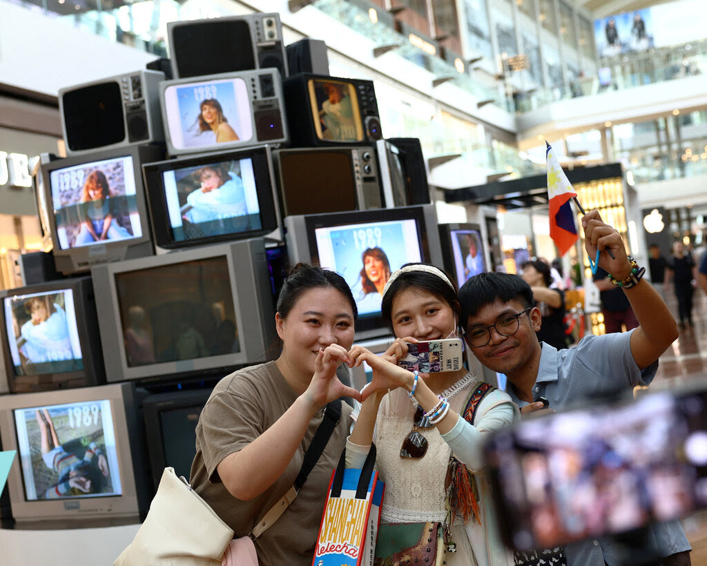 Three people posing for photos in front of many televisions showing Taylor Swift. 