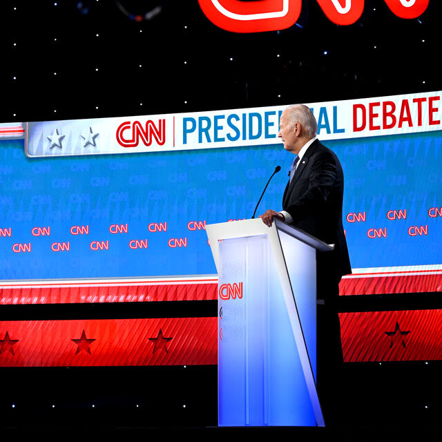 Former President Donald J. Trump and President Biden on a debate stage, each standing at lecterns with microphones. The CNN logo is adorned on the lecterns and on screens behind them.