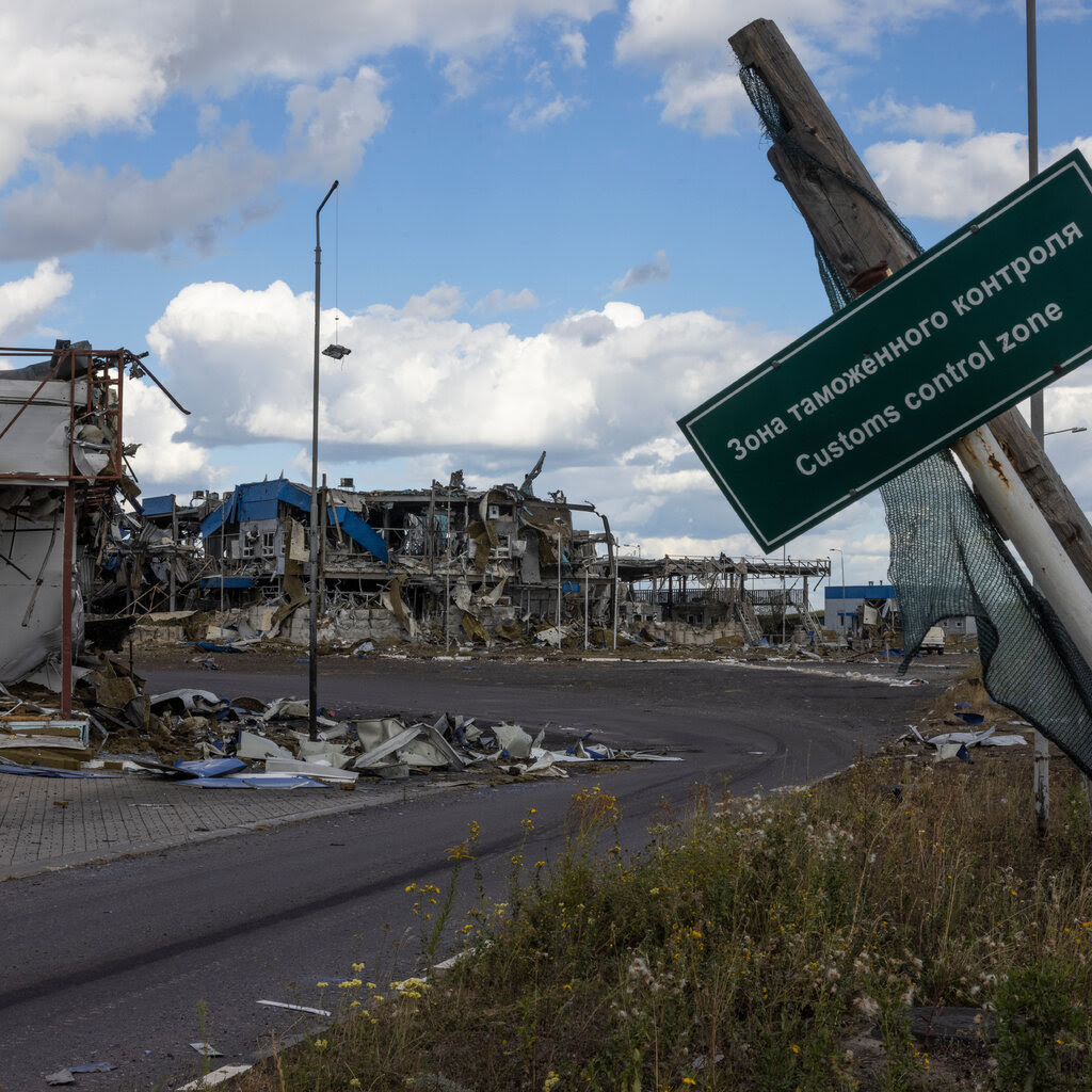 A green sign with white lettering hangs on a tilted post in the forefront, and destroyed buildings are in the rear near the curve of a road.