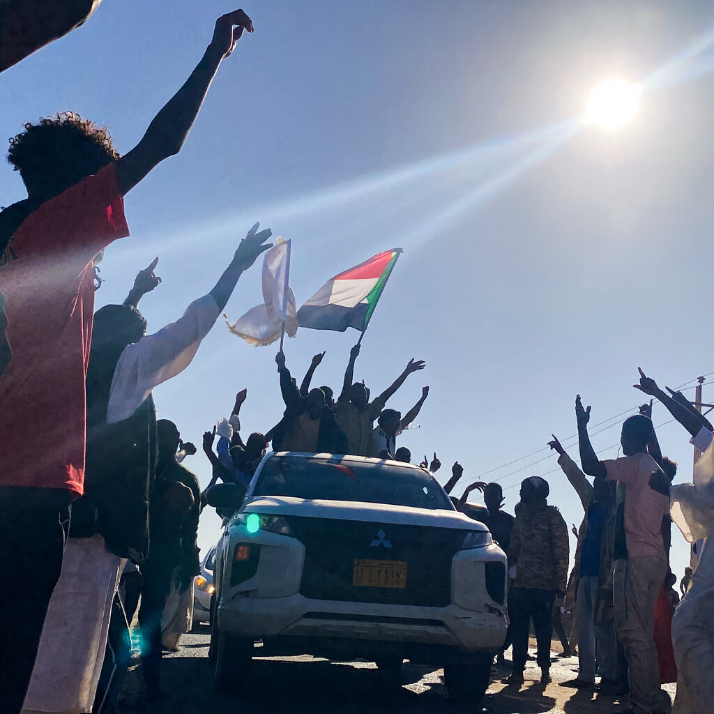 People stand around a car, many of them holding their arms up in celebration.