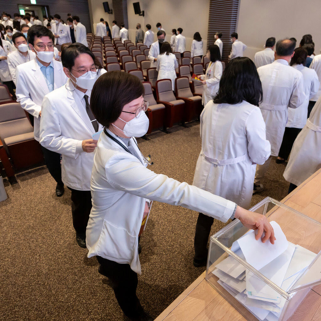 People wearing white jackets walk down the stairs of a lecture hall to place pieces of paper in a clear box. 