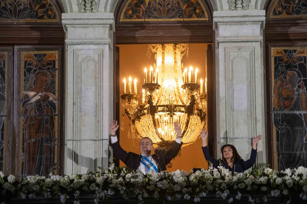 A man and a woman raise their hands in greeting from a flower-bedecked, palatial balcony with an ornate chandelier behind them.