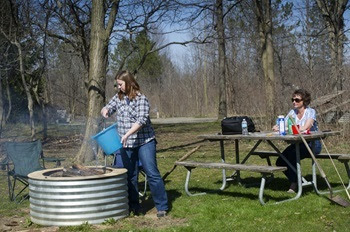 woman in jeans and long-sleeved shirt pours a bucket of water on a campfire as another woman sits at a picnic table next to her