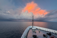 A thunderstorm forms in the sky during sunrise over the bow of a research vessel on open water.