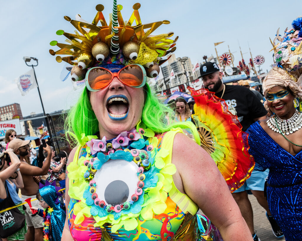 A person in a neon outfit with a big googly eye on their chest. Their mouth is open and they are wearing blue lipstick. 