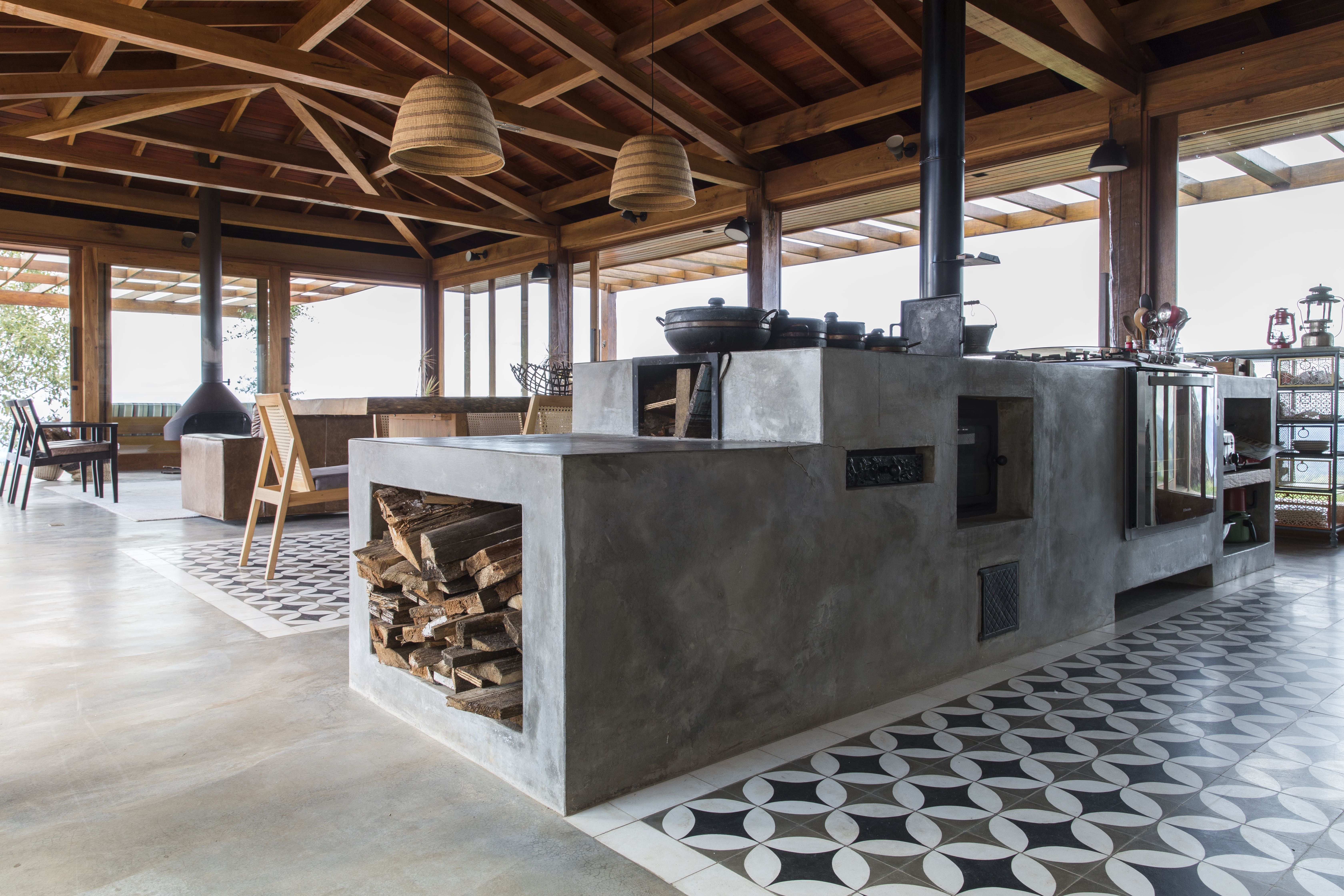 Na sala principal, um fogão à lenha aquece refeições em panelas de barro, destacando-se uma mesa de madeira robusta no espaço de jantar, cercada por ladrilho hidráulico que compõe o tapete desenhada pela Sabella Arquitetura | Foto: Maíra Acayaba 