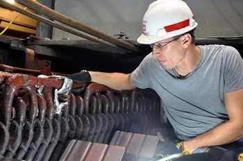 A man in a gray T-shirt, gloves and a white hardhat examines a metal component in a biomass plant