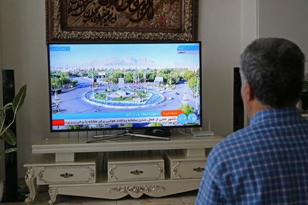 A person in a blue checked shirt watches a television sitting atop an ornate white table.