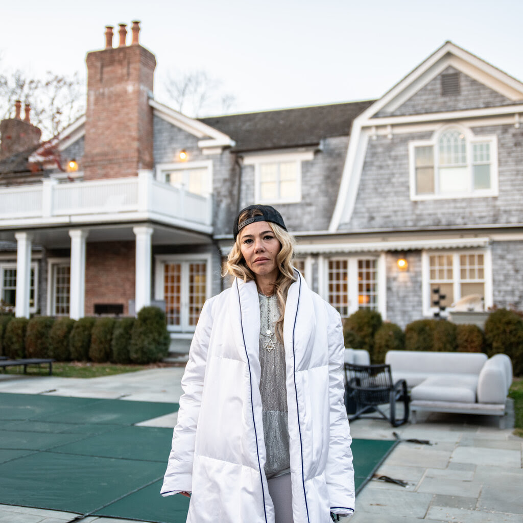 A woman in a winter coat stands outside a home with a prominent fireplace chimney.