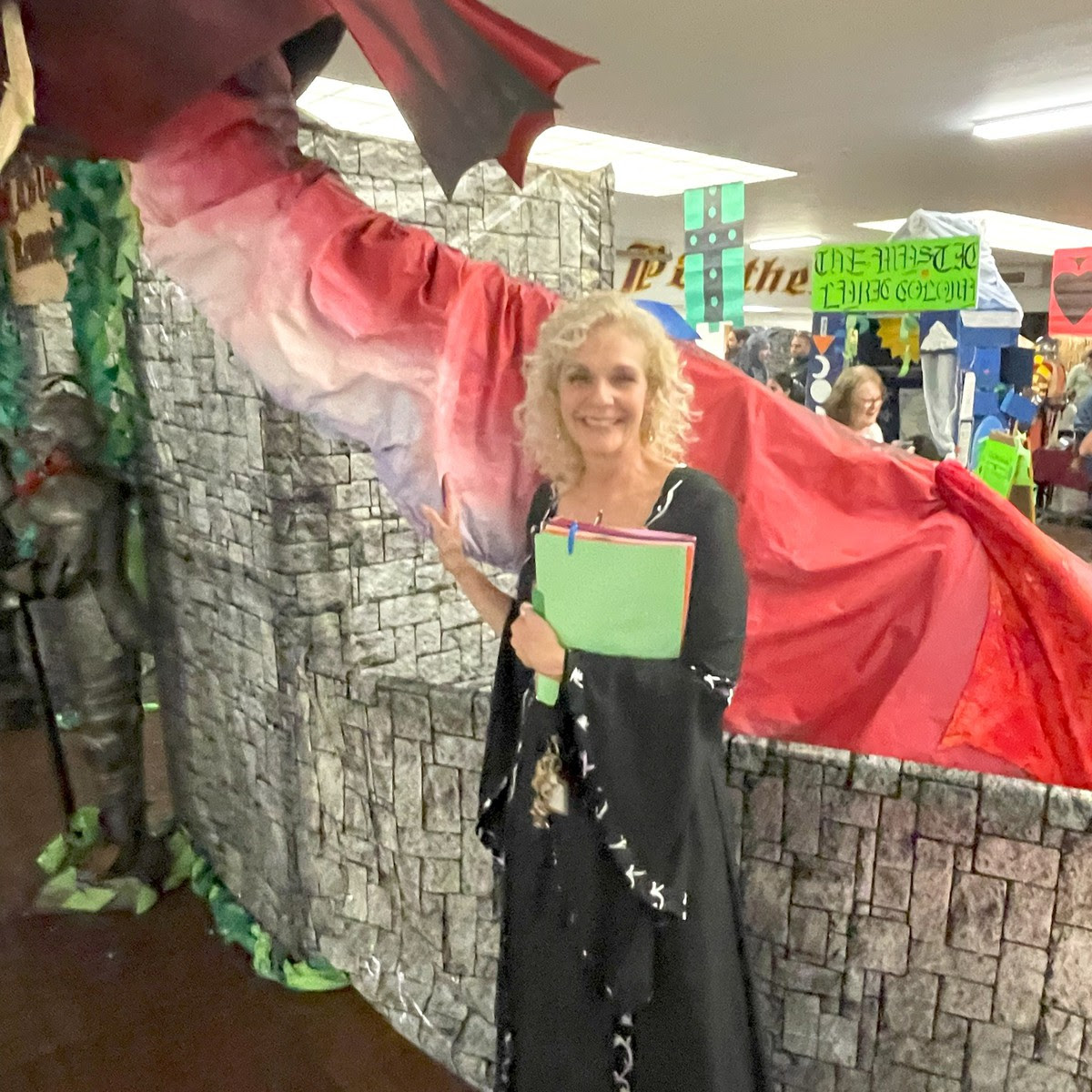 instructor Barbara Sweeney is dressed in a medieval style dress and stands in front of a large paper dragon outside the beautifully painted cardboard "gates" of Castle Kennedy, booths and attendees are visible in the background