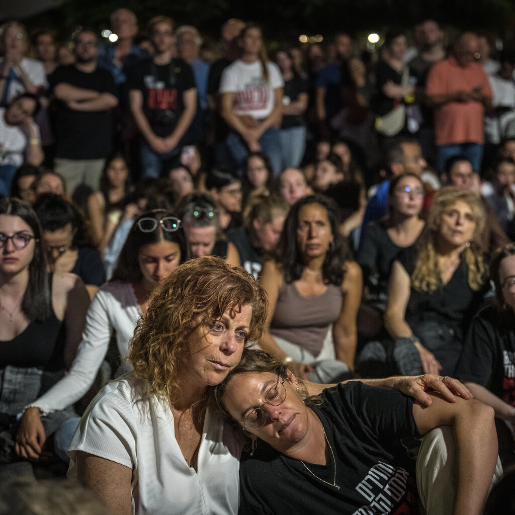 A group of people are sitting on the ground. One woman is leaning her head on another woman's shoulder. More are standing in the background.