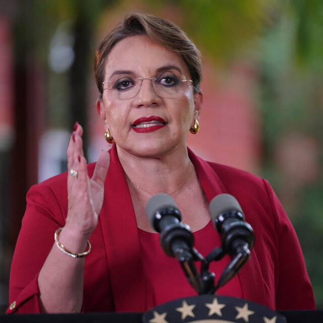 The Honduran president, wearing spectacles and a red outfit, gestures while speaking before two microphones.