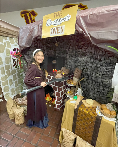 a young lady dressed in medieval clothing interacts inside her booth, made to look like a rustic bakery