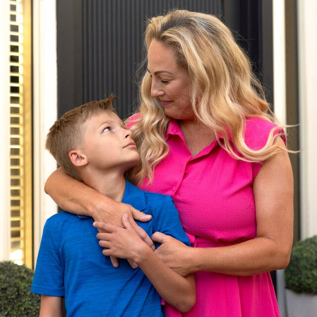 A portrait of Renee Johnson, who wears a bright pink sleeveless dress, and her son, Gage, who wears a blue polo shirt, as they embrace outside the front of a home in Arizona.