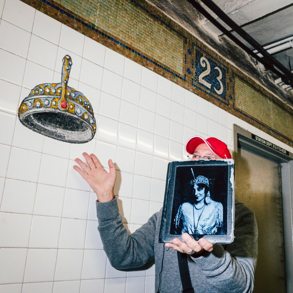 A tour guide holds up an old photo of a woman in elaborate headgear and gestures to a matching mosaic on white tiles inside a subway station.