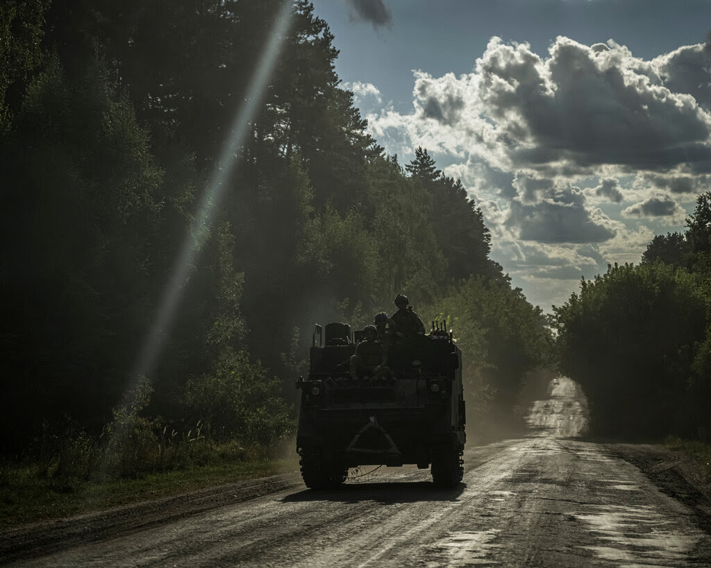 A military vehicle drives down a muddy road with a line of trees on either side. A streak of glare from the sun crosses the image.