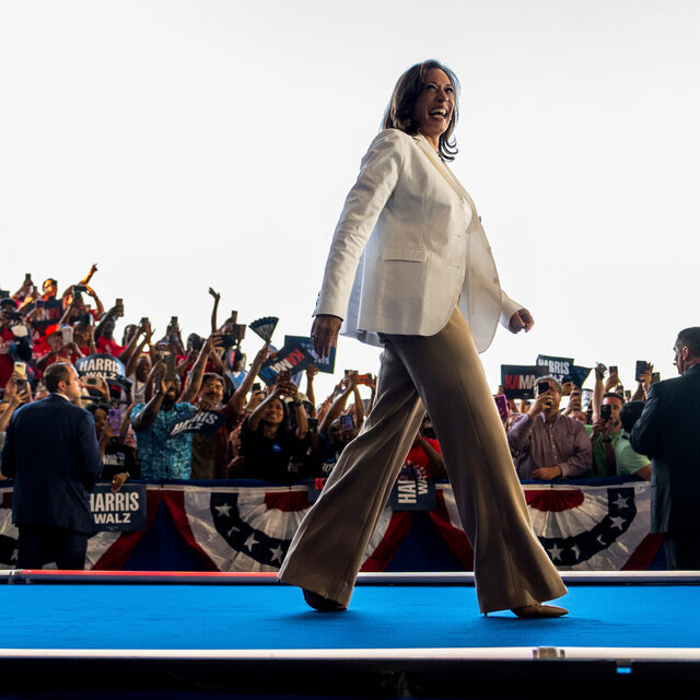 Kamala Harris walking on a blue platform as supporters hold up signs and cheer in the background.