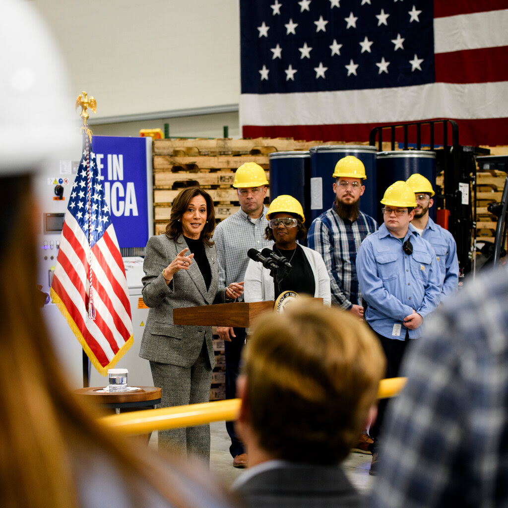 Kamala Harris speaking at a factory with people in yellow hard hats next to her.