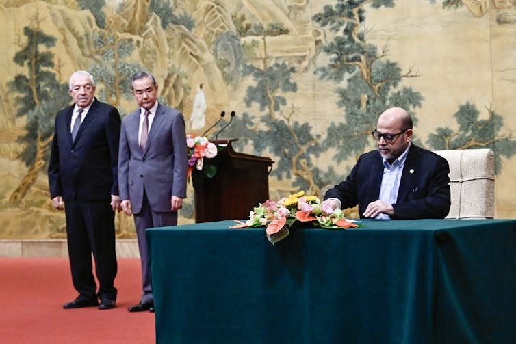 A man at a desk signs a piece of paper as two men standing nearby look on.