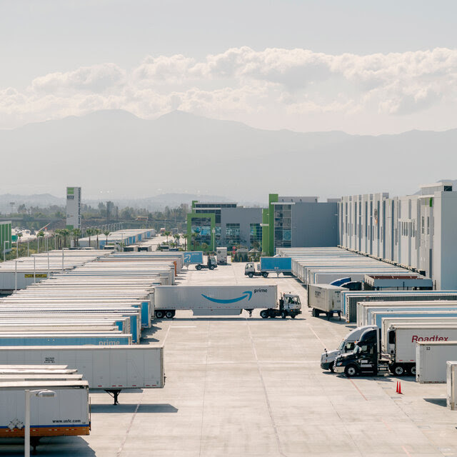 Dozens of semitrailers line a parking lot and loading dock next to a huge warehouse.