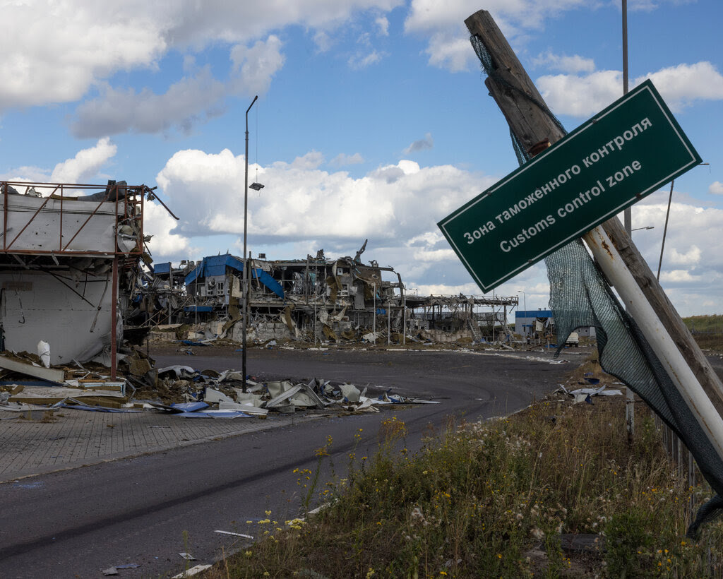 A green sign with white lettering hangs on a tilted post in the forefront, and destroyed buildings are in the rear near the curve of a road.