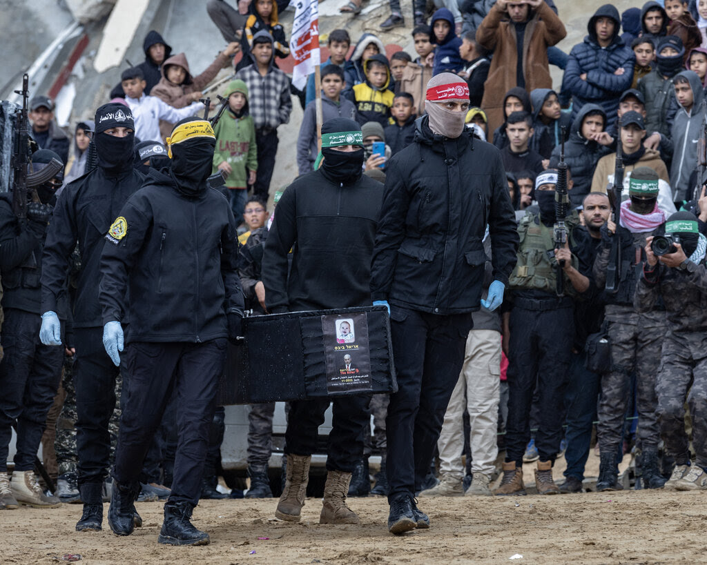 A group of people dressed in black, some with their faces covered, carry a coffin through a crowd.