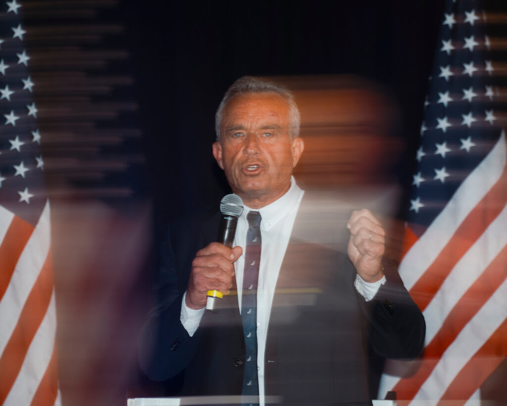 A blurred image of Robert F. Kennedy Jr. speaking into a microphone. 