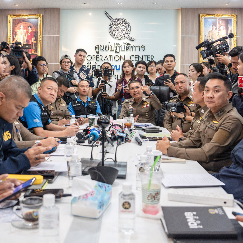 Nearly a dozen officials are seated around a rectangular table, surrounded by photographers and other members of the press. The wall behind them says “Operation Center” on it.