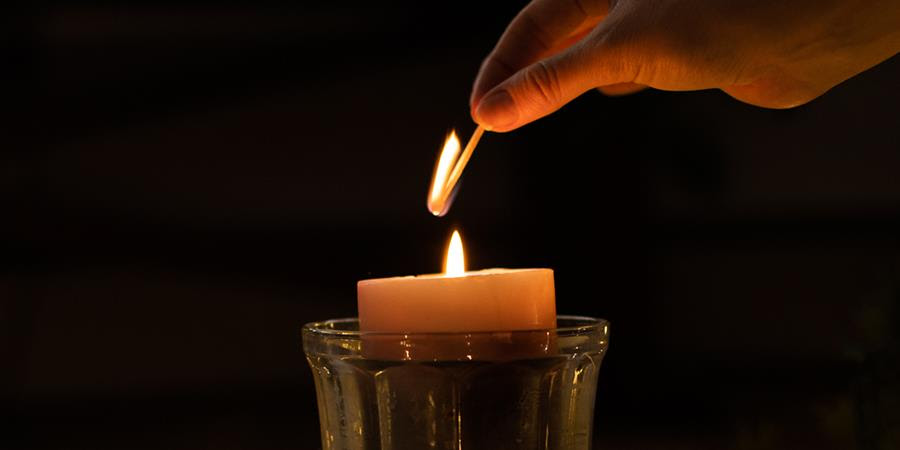 A close up photo of a hand holding a lit match, we see the moment after a candle has been lit against a dark background.