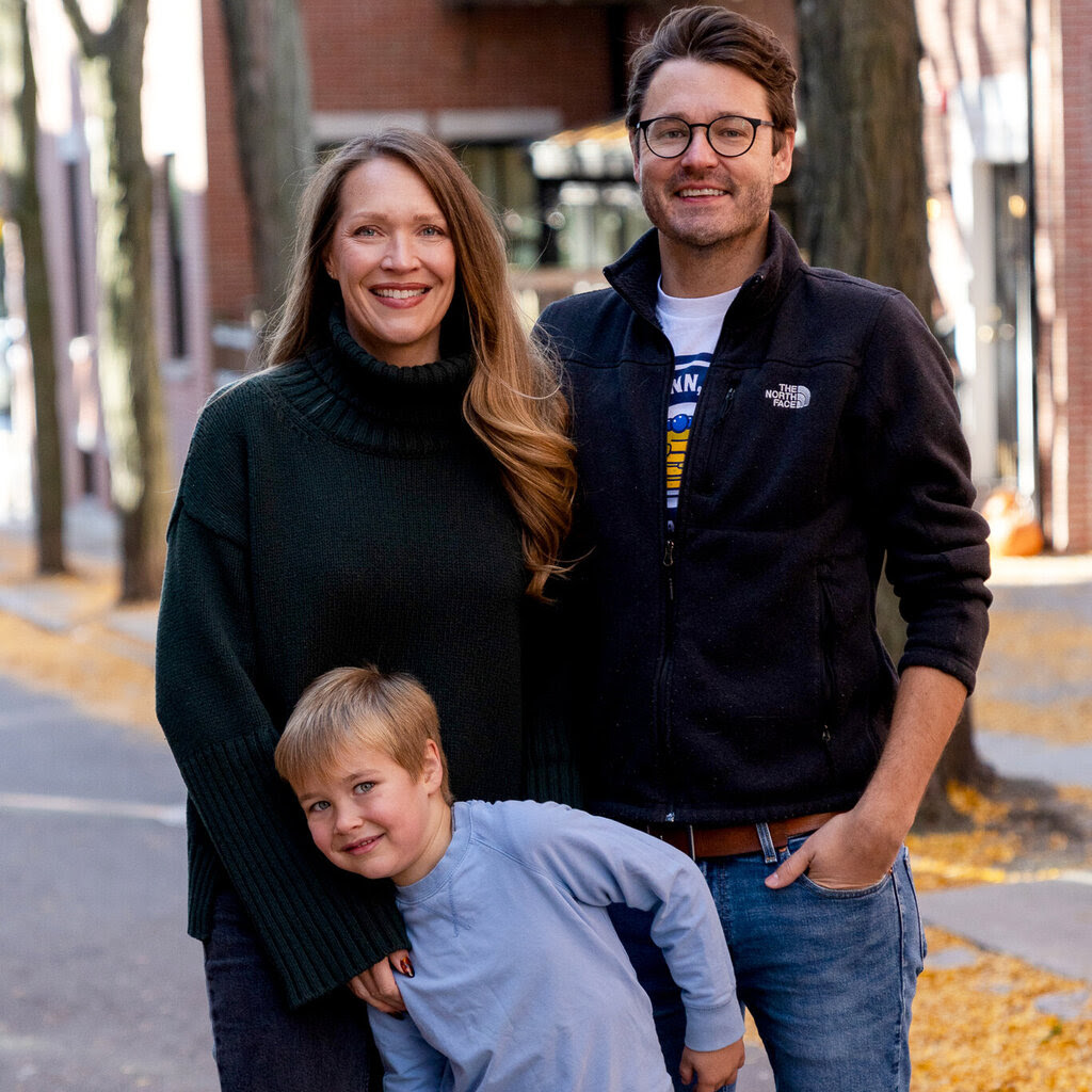 A man in a fleece, woman in a sweater dress and boy in a blue shirt smile on a city street.