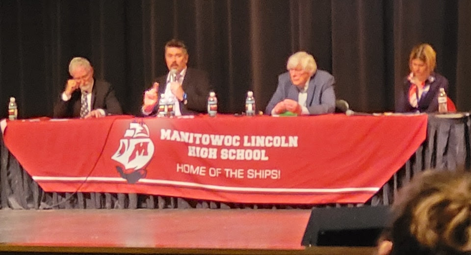 During a recent school board candidates debate, three men and one woman sit at a table, with a banner that reads "Manitowoc Lincoln High School: Home of the Ships!"