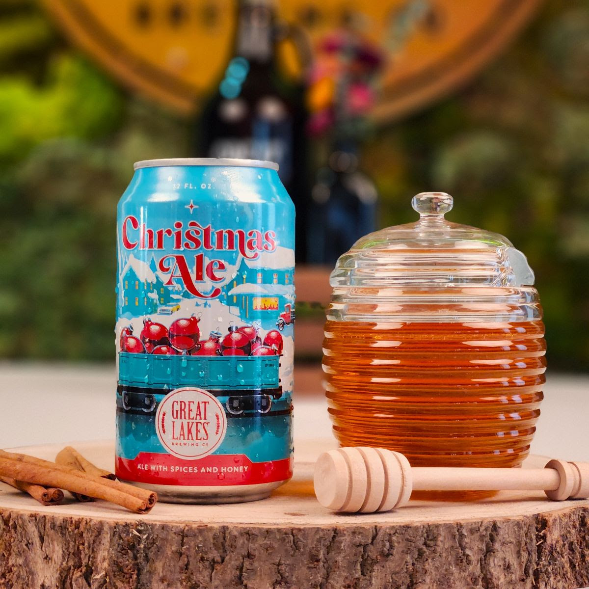 Can of Christmas Ale, a jar of honey with a serving wand, and cinnamon sticks on a log-like wooden stand.