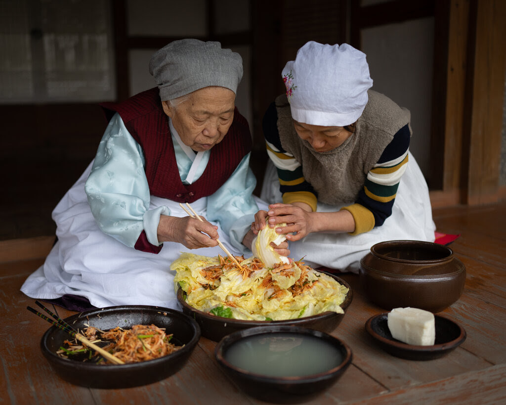 Two women crouch above a large bowl full of cabbage.