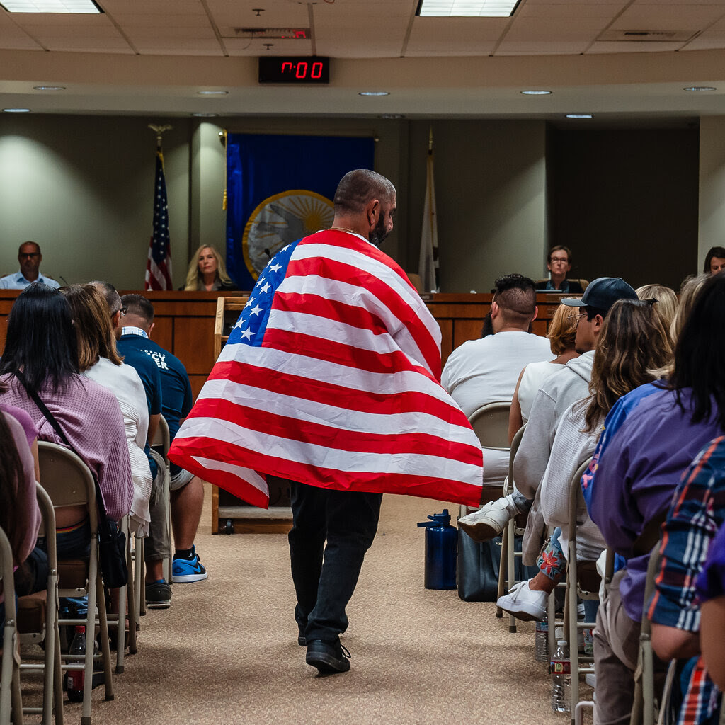 A man wearing an American flag walks down the aisle at a school board meeting. Board members are seated in the background, with spectators seated on each side of the aisle.