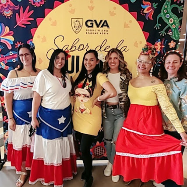  A group of women smiling and posing in front of a colorful backdrop at a GVA event some wearing traditional dresses in bright colors with a banner reading Sabor de la Cultura a joyful and festive atmosphere