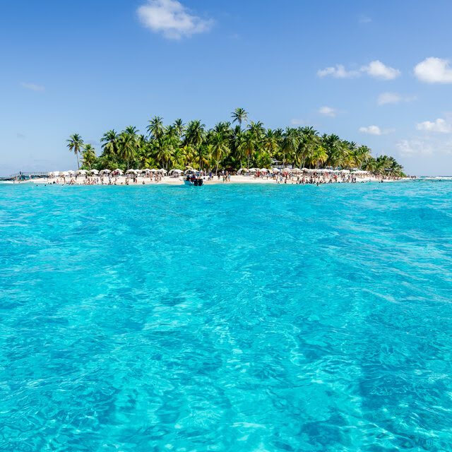 An island with palm trees at its center ringed by beach with beach umbrellas is seen across an expanse of clear blue water. 