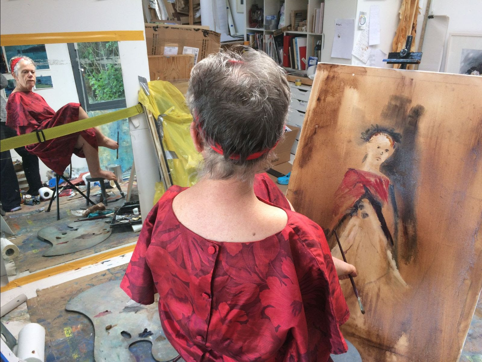 In the foreground, Mary Duffy is seen from behind painting a self-portrait with her left foot, and wearing a red floral-pattern dress. In the background to the left, her reflection is visible, revealing her expression of concentration and the bits and pieces of her artist's studio.