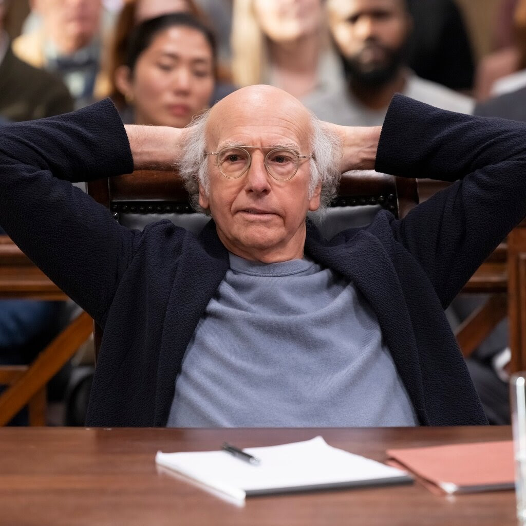 Larry David sits in a courtroom with his hands behind his head