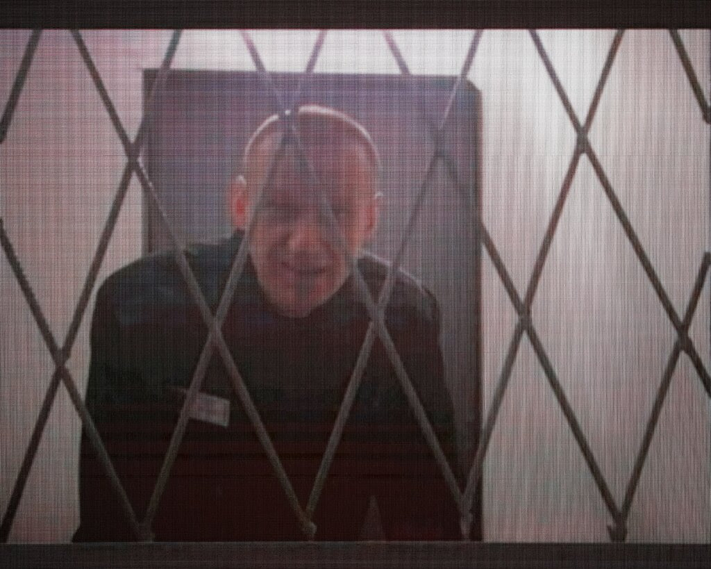 A video still of Aleksei Navalny in black prison garb, seen through a metal grille.