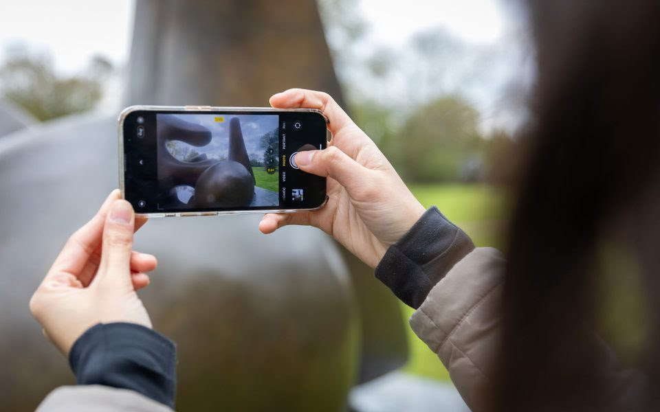 Two hands hold up a smartphone to take a picture of a bronze, abstract sculpture. The artwork is visible on the phone screen.