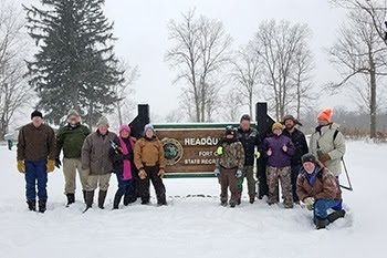 A dozen or so adults in winter coats and gear gather around a brown, rectangular state park entrance sign, with snow all around