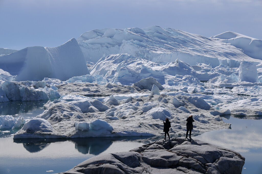 Two people stand on a gray rock on a bright day and look out on melting icebergs.