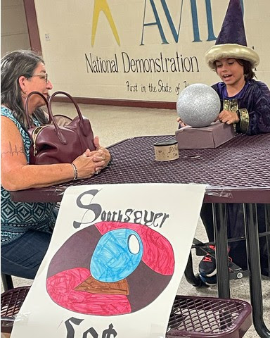 a student sits at a table with purple tablecloth looking into a crystal ball, wears a tall pointed hat and speaks to a guest. The sign on the table reads Soothsayer 50 cents