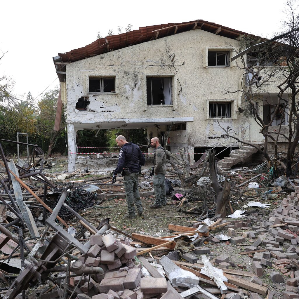 Two men stand in front of a damaged house next to the ruins of a destroyed house.