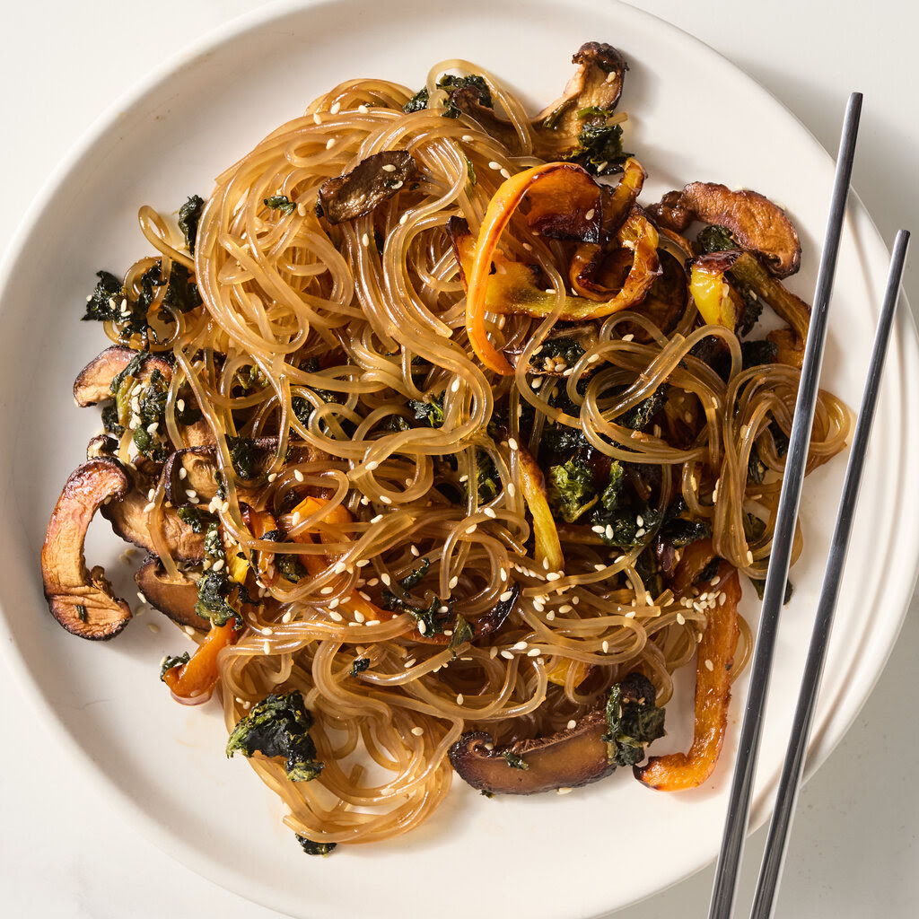 A white plate heaped with glass noodles and slighty-charred roasted vegetables, topped with sesame seeds.